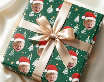 May include: A square gift box wrapped in dark green paper featuring a repeating pattern of white Christmas trees, small yellow stars, and a smiling child wearing a red Santa hat, tied with a wide, smooth, champagne-colored satin ribbon.
