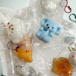 May include: Assortment of colorful, cartoon-like charms in clear plastic packaging. The charms include a light blue puppy, a white cloud, and an orange crescent moon, each with small details and attached keychains.