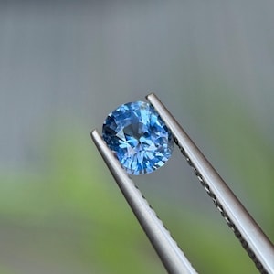 Puede incluir: Una piedra preciosa de zafiro azul con corte cojín sostenida por unas pinzas de metal plateado. La piedra muestra un tono azul aciano vibrante con superficies facetadas que reflejan la luz, creando una apariencia brillante y destellante.
