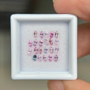 May include: A collection of oval-cut gemstones in various shades of pink and one blue-grey stone, displayed in a white square container. The gemstones are arranged in rows, showcasing their different colors and shapes.