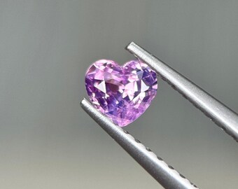 Può includere: Una gemma sfaccettata a forma di cuore, nei toni del rosa e del viola, è tenuta da una pinzetta d'argento. La gemma ha un taglio brillante, che riflette la luce. Lo sfondo è grigio chiaro.
