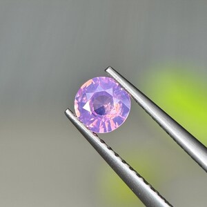 Puede incluir: Una gema redonda y facetada en tonos rosas y morados es sostenida por unas pinzas plateadas. La gema tiene un corte brillante, que refleja la luz y crea un efecto brillante. El fondo es un suave degradado de gris y verde.