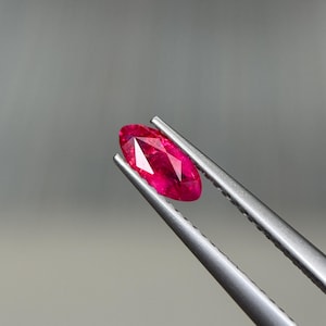 Könnte beinhalten: Ein facettierter, oval geformter Rubin-Edelstein, der von einer silbernen Pinzette gehalten wird. Der Rubin hat eine leuchtende, tiefrote Farbe mit sichtbaren Innenfacetten. Der Hintergrund ist einfarbig grau.