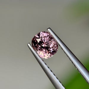 May include: A cushion-cut, pink gemstone is held by a pair of silver tweezers. The gemstone has a faceted surface with varying shades of pink and a subtle sparkle. The background is a soft, blurred gradient of gray and green.