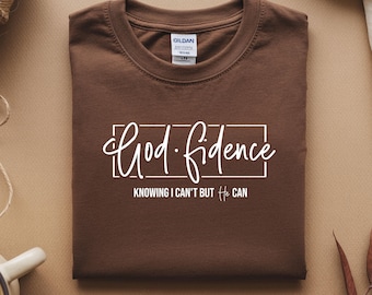 Op de afbeelding: Een gevouwen bruin t-shirt met de woorden "God-fidence" in witte letters, in een witte rechthoek. Daaronder staat "Knowing I can't but He can". Het shirt heeft een ronde hals en korte mouwen.
