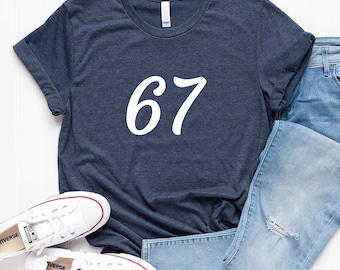 May include: Navy blue t-shirt with the number 67 in white script. The shirt is paired with light blue jeans and white Converse sneakers. The t-shirt has rolled-up sleeves.