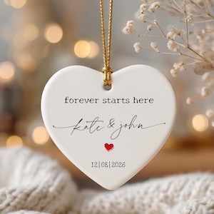 May include: A white heart-shaped ceramic ornament hanging from a gold string. The text reads "forever starts here", "Kate & John", and "12|08|2026" with a small red heart icon centered between the names and the date.