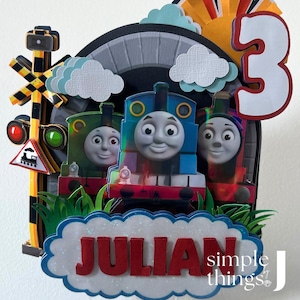 Op de afbeelding: Een gelaagde taarttopper voor een verjaardag met drie cartoon treinen, een spoorwegovergangssignaal en het nummer 3. De naam "JULIAN" staat in rode letters op een wolk-vormige banner. De achtergrond bevat een tunnel en papieren wolken.