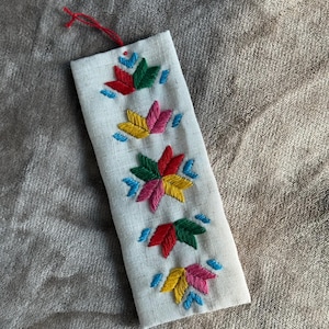 Peut inclure: Marque-page rectangulaire en tissu blanc cassé avec des motifs floraux brodés colorés. La broderie présente des pétales rouges, verts, jaunes et roses avec des accents bleus. Une ficelle rouge est attachée en haut.