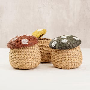 May include: Three woven mushroom-shaped baskets. The baskets have a natural beige color with mushroom caps in brown with white spots, yellow with white spots, and green with white spots. The baskets are made of woven material.