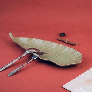 Pode incluir: Uma travessa de cerâmica alongada em forma de folha com um acabamento em degradê verde-claro e creme. Uma colher e um garfo de metal prateado repousam sobre a travessa. Um pau de canela e uma pequena pinha escura estão colocados por perto.