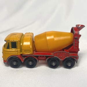 May include: A vintage toy cement mixer truck. The cab is yellow, the mixer drum is orange, and the chassis is red. It has six black wheels and is a classic toy vehicle.