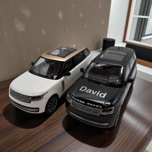 May include: Two toy Range Rover SUVs. One is white with a gold roof, the other is black with the name "David" on the hood. Both have black wheels and a detailed front grille. The cars are displayed on a wooden surface.