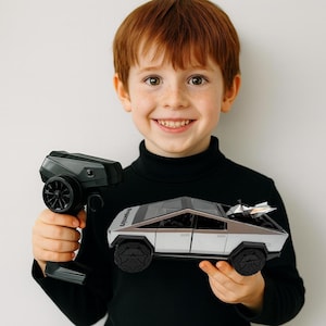 May include: A silver and gray remote-controlled toy car with black wheels and a matching remote control. The toy car has a unique angular design. The child is wearing a black turtleneck.