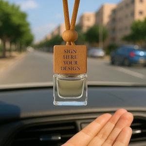 Op de afbeelding: Een auto luchtverfrisser met een houten bovenkant en een doorzichtige glazen fles. De houten bovenkant heeft de tekst "SIGN HERE YOUR DESIGN" gegraveerd. De luchtverfrisser hangt voor een vage achtergrond van een weg en gebouwen.
