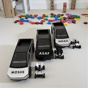 May include: A collection of toy cars, including three black and silver Tesla Cybertruck models with names "MOSHE", "ASAF", and "NOAH" printed on them. Two smaller, futuristic-looking vehicles are also present. Colorful building blocks are scattered in the background.