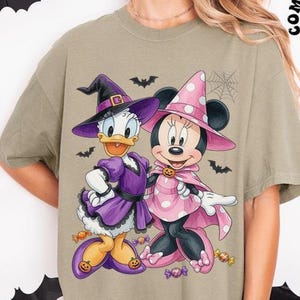 May include: A light olive green t-shirt featuring a graphic print of Daisy Duck and Minnie Mouse dressed in Halloween costumes. Daisy wears a purple witch hat and dress, while Minnie wears a pink polka-dot witch hat and cape with candy.