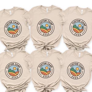 A collection of six beige t-shirts with the same design, featuring a circular logo with a sun, mountains, and text that says "CUSTOM CAMP" and "2025".