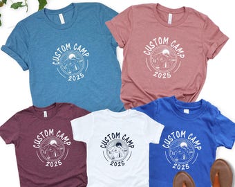May include: A collection of short-sleeved t-shirts in various colors, including blue, red, white, and burgundy. Each shirt features a white circular graphic with the text "CUSTOM CAMP 2025" and a mountain and tent design.