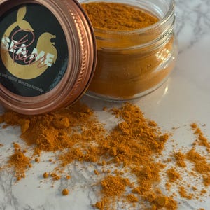 May include: A clear glass jar filled with bright orange powder, with a copper-colored lid resting beside it. The lid features a black label with gold text and a mermaid tail and lion graphic. The text on the label reads "SEA ME" and "and natural skin care remedy."