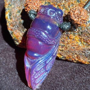 Può includere: Ciondolo viola e blu a forma di cicala. Il ciondolo traslucido presenta dettagli intricati ed è sospeso a un cordino nero con perline decorative. Gioielli fatti a mano.