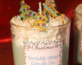 Puede incluir: Una vela de vidrio transparente con una base de cera verde claro, cubierta con cera batida blanca y purpurina dorada. La vela presenta tres adornos decorativos de árbol de Navidad. La etiqueta dice "Oh Christmas Tree" y "Sprinkle Glow Candle Co."