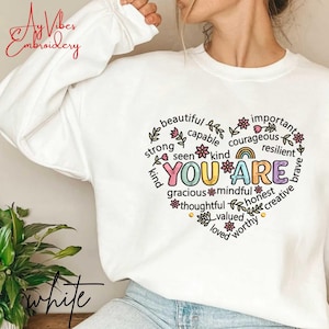 Puede incluir: Sudadera blanca con un diseño en forma de corazón. El diseño incluye las palabras "YOU ARE" en letras coloridas, rodeadas de afirmaciones positivas como "beautiful", "strong" y "kind". La sudadera también tiene el texto "Ay Vibes Embroidery" y "white".