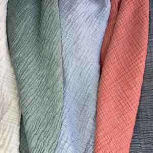 May include: Close-up of several fabrics in various colors. The fabrics include white, sage green, light blue, coral, and navy blue. The material has a textured, crinkled appearance, suggesting a lightweight fabric like gauze or muslin.