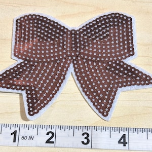 May include: Brown sequin bow with white polka dots and a white border. The bow is approximately 4 inches wide. The bow is on a light wood surface.