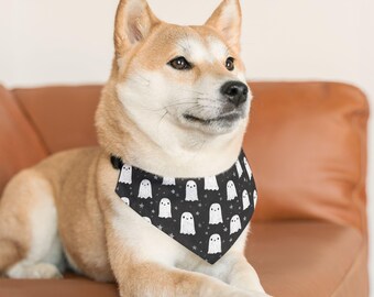 May include: A Shiba Inu dog wearing a black bandana with a white ghost and star pattern. The dog is lying on a brown leather couch. The bandana is tied around the dog's neck.