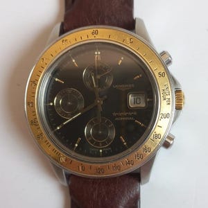 Op de afbeelding: Een Longines Admiral polshorloge met een donkere wijzerplaat, een goudkleurige lunette en een bruine leren band. Het horloge heeft subwijzerplaten, een datumweergave en de tekst "Admiral". De lunette heeft markeringen voor eenheden per uur.