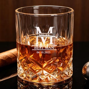 Puede incluir: Un vaso transparente lleno de líquido ámbar, con un patrón de diamante tallado y texto grabado en blanco que dice MILES, BEST MAN, 06.10.2025. Un cigarro marrón y una pequeña esfera de metal plateado están colocados junto al vaso.