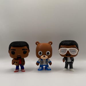Puede incluir: Tres figuras de Funko Pop. Una figura es marrón con una chaqueta marrón, pantalones azules y zapatos rojos, sosteniendo un osito de peluche. La segunda figura es un oso marrón con un atuendo azul y gris. La tercera figura lleva gafas de sol y un traje negro.