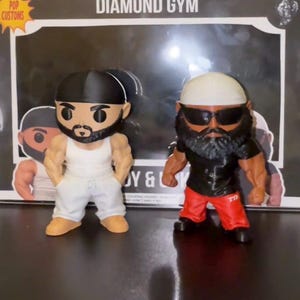May include: Two custom-made Diamond Gym figurines. One figure wears a white tank top and gray sweatpants, the other wears a black shirt and red pants. Both have muscular builds, beards, and head coverings. The background reads "Diamond Gym".