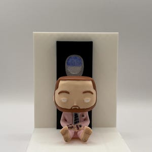 May include: A figurine of a seated person with a beard, wearing a pink suit, holding a game controller. The figurine is on a white base with a black rectangle and a blue and white oval shape above it.