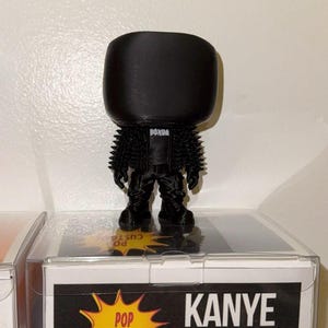 Puede incluir: Figura de vinilo negra de Kanye West con una chaqueta puntiaguda y la palabra "DONDA" en el pecho. La figura se muestra en una caja de plástico transparente con las palabras "POP CUSTOMS" y "KANYE WEST" en la caja.