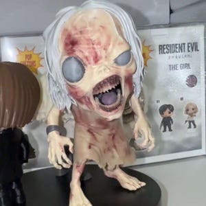 Può includere: Statuetta dettagliata di un personaggio simile a uno zombie con carnagione pallida, bocca aperta e denti scoperti. La figura ha i capelli bianchi e indossa abiti laceri. La figura è su una base nera, con una scatola sullo sfondo che dice "RESIDENT EVIL".