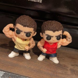 以下が含まれることがあります： 上腕二頭筋を曲げている筋肉質な男性の2つのFunko Popフィギュア。1つは黄色のタンクトップと黒のショーツ、もう1つは赤いタンクトップと青のショーツを着ています。どちらも腕にタトゥーがあり、白いスニーカーを履いています。