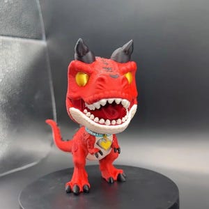 Pode incluir: Uma figura de dinossauro vermelha com uma cabeça grande, olhos amarelos e chifres pretos. A criatura tem uma mandíbula branca com dentes visíveis, uma coleira azul-clara e um pingente amarelo em forma de coração. Fica em duas patas com uma cauda.