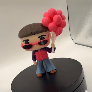 May include: A stylized vinyl figure with a bowl-cut hairstyle, wearing pink-rimmed sunglasses, a red and dark blue jacket, and blue pants. The figure holds a cluster of bright pink balloons attached to white strings in one hand.