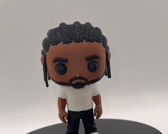 May include: A vinyl figure of a person with dark skin, wearing a white t-shirt, black pants, and white sneakers. The figure has black braids and a small beard. It stands on a black circular base.