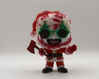May include: A figurine of a Santa Claus figure with a menacing expression. The figure wears a red Santa suit, a blood-splattered hat, and green Christmas tree glasses. It holds a small axe and has a belt with a square buckle.