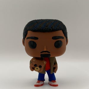Puede incluir: Una figura de vinilo de una persona con piel oscura, cabello negro y un peque&ntilde;o bigote. La figura lleva una camisa roja, pantalones azules, una chaqueta marr&oacute;n y zapatillas rojas y blancas. La figura sostiene un peque&ntilde;o oso de peluche.