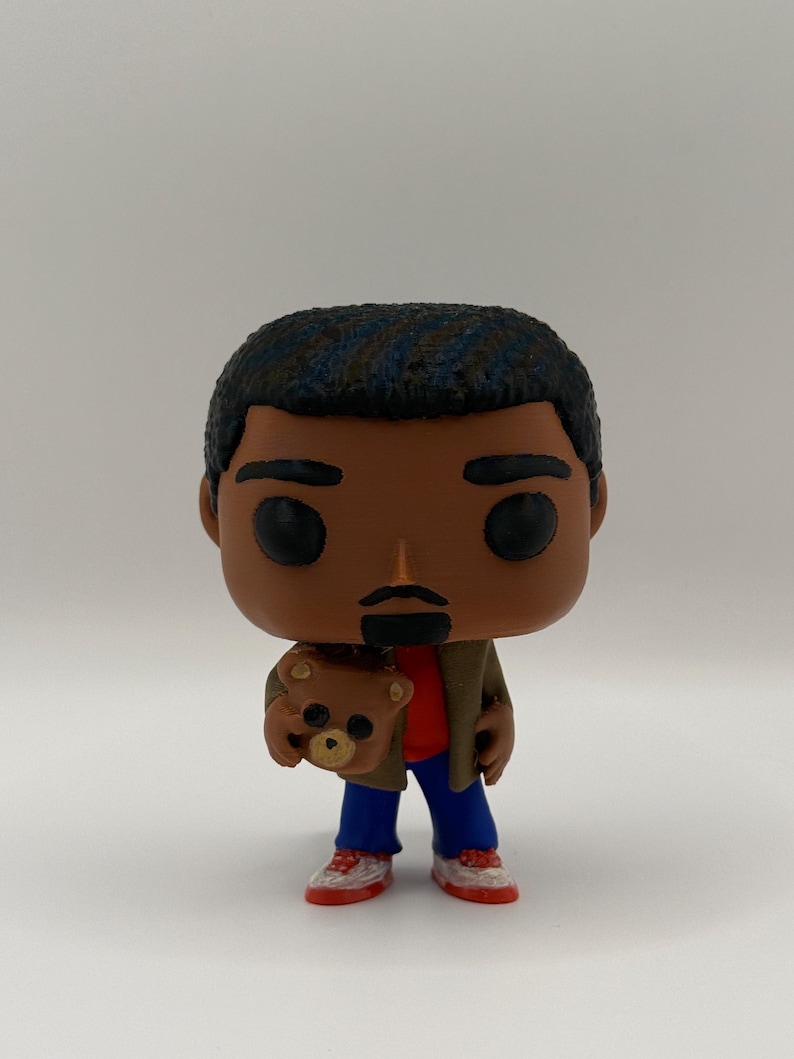 Puede incluir: Una figura de vinilo de una persona con piel oscura, cabello negro y un peque&ntilde;o bigote. La figura lleva una camisa roja, pantalones azules, una chaqueta marr&oacute;n y zapatillas rojas y blancas. La figura sostiene un peque&ntilde;o oso de peluche.
