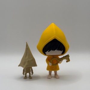 Könnte beinhalten: Zwei Figuren: eine in einem gelben Kapuzenmantel, der einen Schlüssel hält, die andere in einem beigefarbenen Kegelhut. Die gelbe Figur hat schwarzes Haar und ein gelbes Outfit. Die beige Figur hat einen Kegelhut. Beide auf weißem Hintergrund.