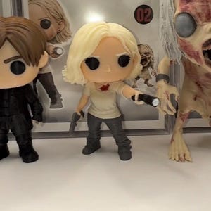 Puede incluir: Colección de figuras de vinilo de la serie de televisión The Walking Dead. Las figuras incluyen una mujer rubia con una pistola y una linterna, un zombi y un hombre de negro. Las figuras están en una vitrina.