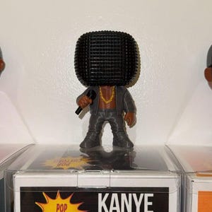 Puede incluir: Una figura de vinilo Funko Pop de Kanye West, con un traje gris y un micrófono. La figura tiene una cabeza negra cuadrada y una cadena dorada. La caja de abajo dice "Kanye West" y "Pop Customs".