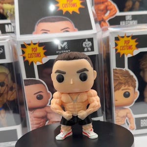 May include: A custom Pop! vinyl figure of a muscular man, with a tan complexion, black shorts, and white trainers with red accents. The figure is on a black display stand. The packaging behind the figure has the text "POP CUSTOMS".