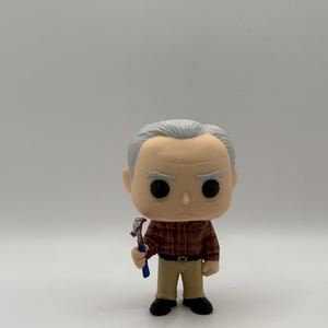 Op de afbeelding: Een Funko Pop vinyl figuur van een man met grijs haar, gekleed in een bruin geruit overhemd, een beige broek en zwarte schoenen. Hij houdt een blauwe en rode bijl vast. De figuur heeft grote zwarte ogen en een neutrale uitdrukking.