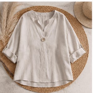 May include: A beige linen-blend blouse featuring a V-neckline, a single central button closure, and three-quarter length sleeves with folded cuffs. The garment has a relaxed, loose-fitting silhouette with a straight hemline and visible fabric texture.
